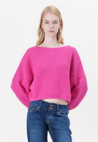 Pull en crochet de couleur rose vif avec une coupe oversize, un large col et de longues manches, associé à un jean bleu doté de trois boutons.
