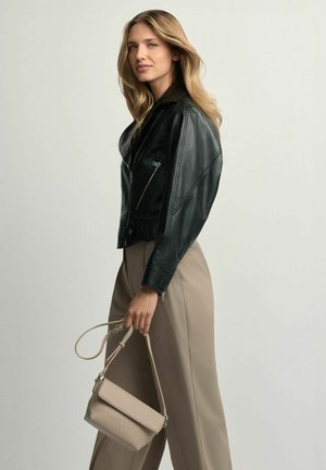 Kobieta z długimi blond włosami, ubrana w czarną skórzaną kurtkę i beżowe spodnie, trzymająca beżową torebkę za pasek.