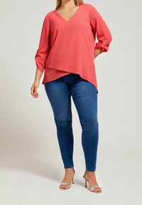 Top corallo a V con maniche a tre quarti, design a strati; abbinato a jeans skinny blu scuro e décolleté argentate con cinturini.