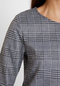 Mustikka- ja valkoisella houndstooth-kuvioidulla paidalla, jossa on pyöreä pääntie ja pitkät hihat. Paita on valmistettu teksturoidusta kankaasta ja siinä on jämäkkä muotoilu.
