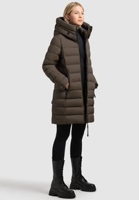 Ciemnobrązowa puchowa kurtka z wysokim kołnierzem, zamkiem z przodu, bocznymi kieszeniami i ściągaczami na mankietach, noszona na czarnych legginsach i glanach.