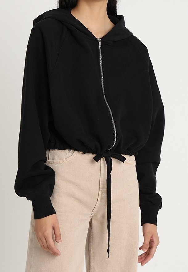 Femme portant un sweat à capuche court noir zippé avec cordon à la taille et un pantalon beige taille haute sur un fond clair uni.