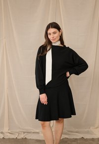 Moteris dėvi juodą ilgomis rankovėmis suknelę ir baltą šaliką ant kaklo, stovinti priešais smėlio spalvos audinio foną.