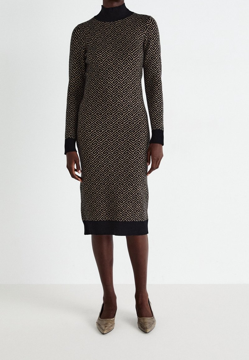 Femme portant une robe-pull midi ajustée à motifs géométriques noirs et beiges, manches longues avec poignets noirs, associée à des escarpins beiges à bout pointu.