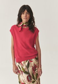 Top in magenta a maniche corte, abbinato a una gonna a fiori con foglie verdi e fiori rossi. Accento in bracciale d'oro.