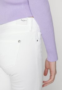 Jeans blancos con una textura suave, que presentan una sutil etiqueta de logo. Combinados con un suéter de color púrpura claro, de canalé y con mangas ajustadas.