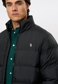 Must must jacket kõrge kaelusega, millel on tähelepandamatud horisontaalsed õmblused ja väike hõbedane logo vasakul rinnal.