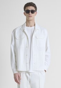 Chaqueta blanca de botones delanteros que cuenta con dos bolsillos en el pecho, cuello redondeado y una textura suave, llevada sobre una camiseta blanca.