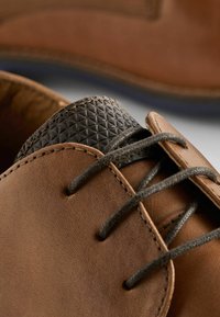 Zapatos de cuero marrón con un acento negro texturizado, que presentan un acabado mate y suave, cordones redondos y detalles de costura visibles.