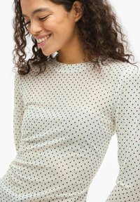 Długi rękaw, top wykonany z prześwitującego materiału z czarnymi kropkami na jasnym tle. Posiada okrągły dekolt i dopasowaną sylwetkę.