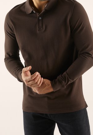 Brązowa polo z długim rękawem, wykonana z miękkiego materiału, z zapinanym kołnierzykiem i wąskim krojem. Teksturowane mankiety i małe rozcięcie po boku.