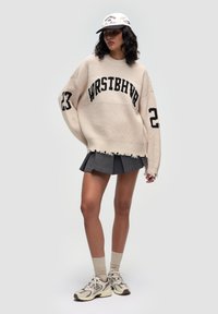 Beige Oversized-Pullover mit schwarzem "WRSTBHVR"-Logo und der Nummer 23. Kombiniert mit einem grauen Faltenrock, beigen Knöchelsocken und Sneakers in neutralen Tönen.