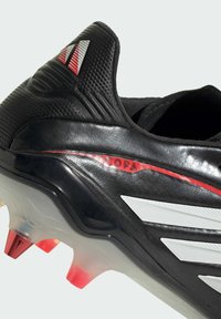 Zwarte voetbalschoen met een gestructureerd bovenwerk, witte strepen en rode accenten. Transparante zool met roodpuntige noppen. Gemarkeerd met "COPA".