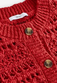 Gilet en tricot rouge avec un design en maille ouverte, deux grands boutons beiges et une étiquette de marque blanche avec "NEXT" brodée dessus.