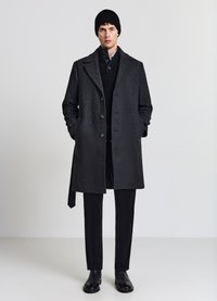 Cappotto di lana nero con finitura testurizzata, caratterizzato da chiusura frontale con bottoni, tasche laterali e un berretto di maglia nero coordinato.