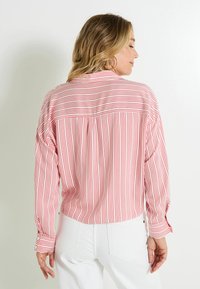 Camisa de manga larga a rayas rojas y blancas con un corte relajado, que presenta un yugo dividido en la parte posterior y puños abotonados, combinada con unos jeans blancos.