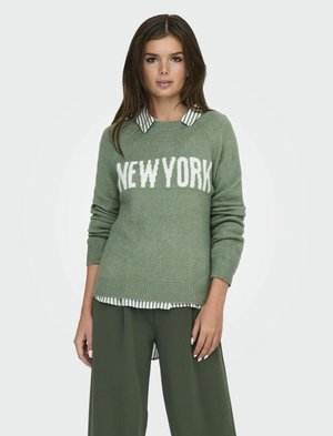 Maglione verde a maglia con "NEW YORK" in bianco. Indossato sopra una camicia a righe con colletto. Abbinato a pantaloni verdi larghi. Tessuto morbido e vestibilità rilassata.