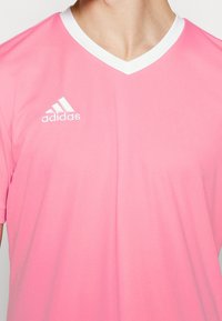 Rosa träningsskjorta med V-ringning, korta ärmar och vit krage. Har en vit Adidas-logotyp på vänster bröst. Slät textur.