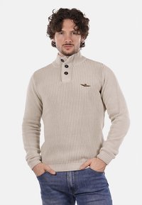 Pullover beige lavorato a maglia con colletto alto, tre bottoni scuri e un piccolo logo marrone sul lato sinistro del petto. Presenta polsini e orlo a costine.