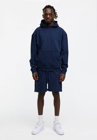 Dunkelblaue Kapuzenjacke mit Fronttasche und passenden Shorts, aus weichem Material, kombiniert mit weißen Turnschuhen und Knöchelsocken.