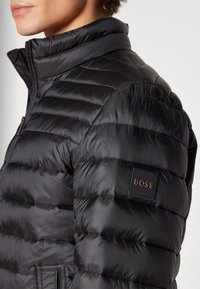 Giubbotto imbottito nero con texture trapuntata, colletto alto e patch con logo sulla manica, con chiusura frontale con zip. Materiale liscio.