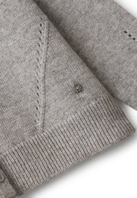 Světle šedý pletený kardigan s žebrovanými manžetami a lemem, který má na rukávu poblíž zápěstí malou kovovou nášivku s logem.