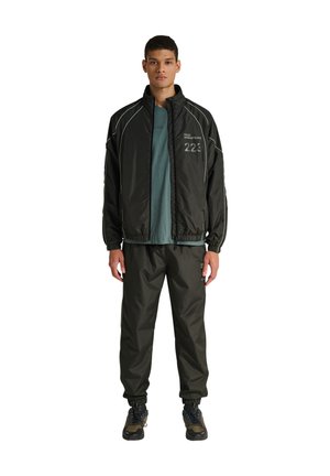 Jonge man staand, gekleed in een zwart sportjack en broek met witte biezen, een groenblauw T-shirt en zwarte sportschoenen tegen een witte achtergrond.