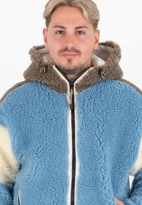 Lichtblauwe fleece jas met een bruine capuchon en accenten, voorzien van een rits aan de voorkant en een textuurstof over de hele jas.