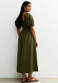 Olijfgroen maxi-jurk met pofmouwen, een gesmokte lijfje en een vloeiende rok. Draagt met bruine sandaaltjes met bandjes, gezien van de achterkant.