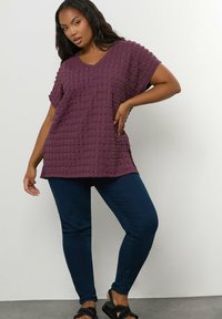 Fioletowa bluzka o fakturze, z dekoltem w serek i krótkimi rękawami, zestawiona z ciemnoniebieskimi dopasowanymi dżinsami i czarnymi sandałami. Tło z płaską powierzchnią.
