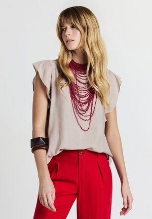 Blusa beige a maniche corte con spalle arricciate e una collana rossa a strati, abbinata a pantaloni rossi a vita alta e a un braccialetto scuro.
