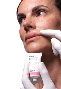 Tubetto bianco di "Instant Anti-Wrinkle Lip Contour" con accenti rosa, tenuto vicino a un volto. La pelle appare liscia, indossando guanti bianchi.