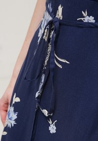 Marineblauwe stof met een lichtblauw en wit bloemmotief, vastgebonden met een smalle strik op de taille, en een hand die ernaast rust.