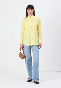Camicia gialla con colletto e tasca, abbinata a jeans a gamba larga blu chiaro. Una piccola borsa marrone e ballerine nere completano il look.