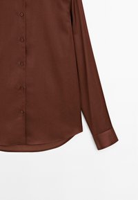 Chemise à manches longues de couleur marron avec une texture lisse, fermeture à boutons sur le devant et ourlet arrondi. Présente des boutons assortis et des détails aux poignets.