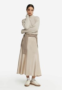 Jasno beżowy sweter dzianinowy z wysokim kołnierzem, zestawiony z satynową midi spódnicą w delikatnym beżu oraz białymi sneakersami z brązowymi akcentami.