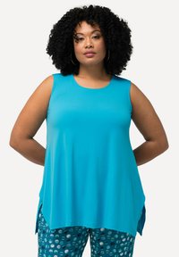 Una camiseta sin mangas color teal con un escote redondo, que presenta un corte suelto y fluido con aberturas laterales para mayor comodidad. Combinada con pantalones azules con estampado.