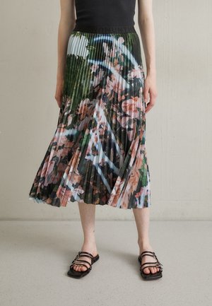 Kobieta w plisowanej, kwiecistej spódnicy midi i czarnym bezrękawnym topie, stojąca na jasno nakrapianej podłodze w sandałach.