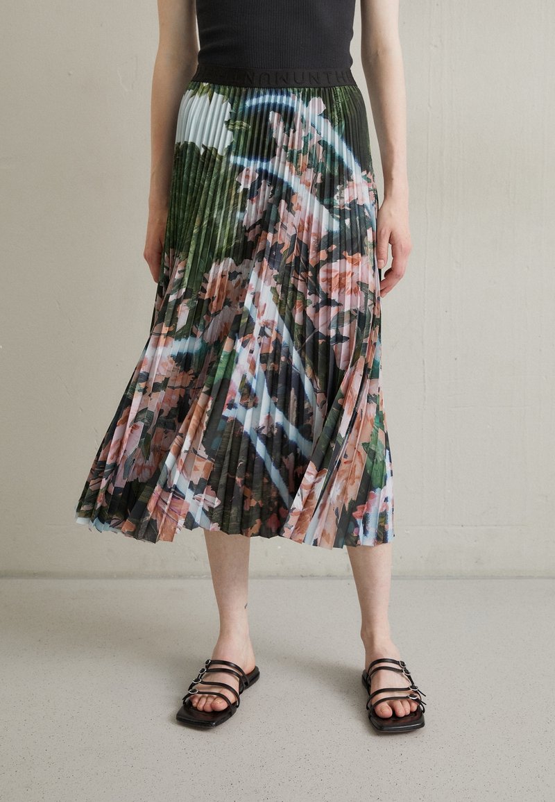 Femme portant une jupe midi plissée à fleurs et un top noir sans manches, debout sur un sol clair tacheté avec des sandales.