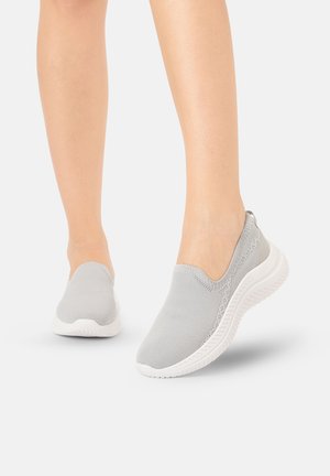 Voeten die lichtgrijze gebreide slip-on sneakers met witte gestructureerde zolen dragen, lopend op een effen witte achtergrond.