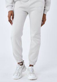 Joggers gris claro hechos de una tela suave; cuentan con puños elásticos y un logo sencillo. Combinados con zapatillas blancas voluminosas.