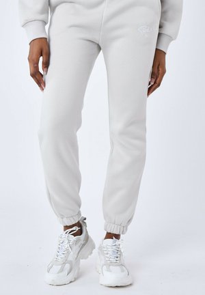 Joggeurs gris clair en tissu doux ; avec des poignets élastiques et un logo simple. Associés à des baskets blanches volumineuses.