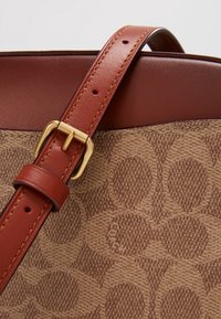 Ruskea kangaslaukku, jossa on teksturoitu kuvio, täydentää sileä burgundinpunainen remmi ja kullanväriset metalliosat.