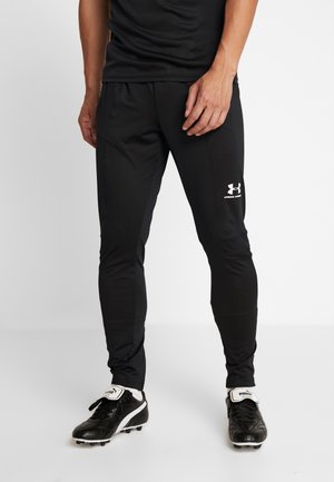 Mężczyzna w czarnych sportowych spodniach Under Armour i czarnych butach piłkarskich Puma stoi na białym tle.