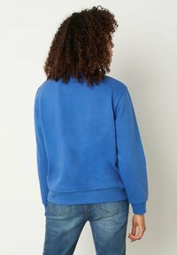 Felpa blu con scollatura rotonda, maniche lunghe e polsini a costine. Tessuto liscio senza motivi o accenti visibili, abbinata a jeans in denim.