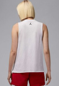 Camiseta blanca sin mangas con cuello redondo, que presenta un logo negro de Jumpman en la parte posterior. Combinada con pantalones cortos rojos, mostrando un look atlético y casual.