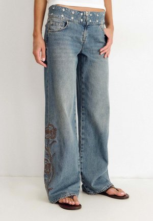 Mujer con vaqueros azul claro de pierna ancha con bordado floral en la parte inferior de la pierna y cintura con tachuelas, combinados con sandalias marrones.