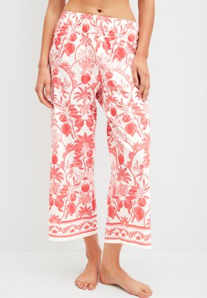 Vrouw die witte, wijd uitlopende broek draagt met een koraalroze bloemen- en fruitprint, blootsvoets staand tegen een effen lichte achtergrond.