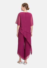 Combinaison en chiffon violet avec des manches courtes et fluides, ourlet asymétrique et pantalons larges. Fermeture à glissière au dos. Sandales à talons bloc argentés.