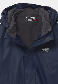 Navy waterproof jacket with a gray fleece lining, zipper closure, and a textured logo patch featuring five circular cutouts.